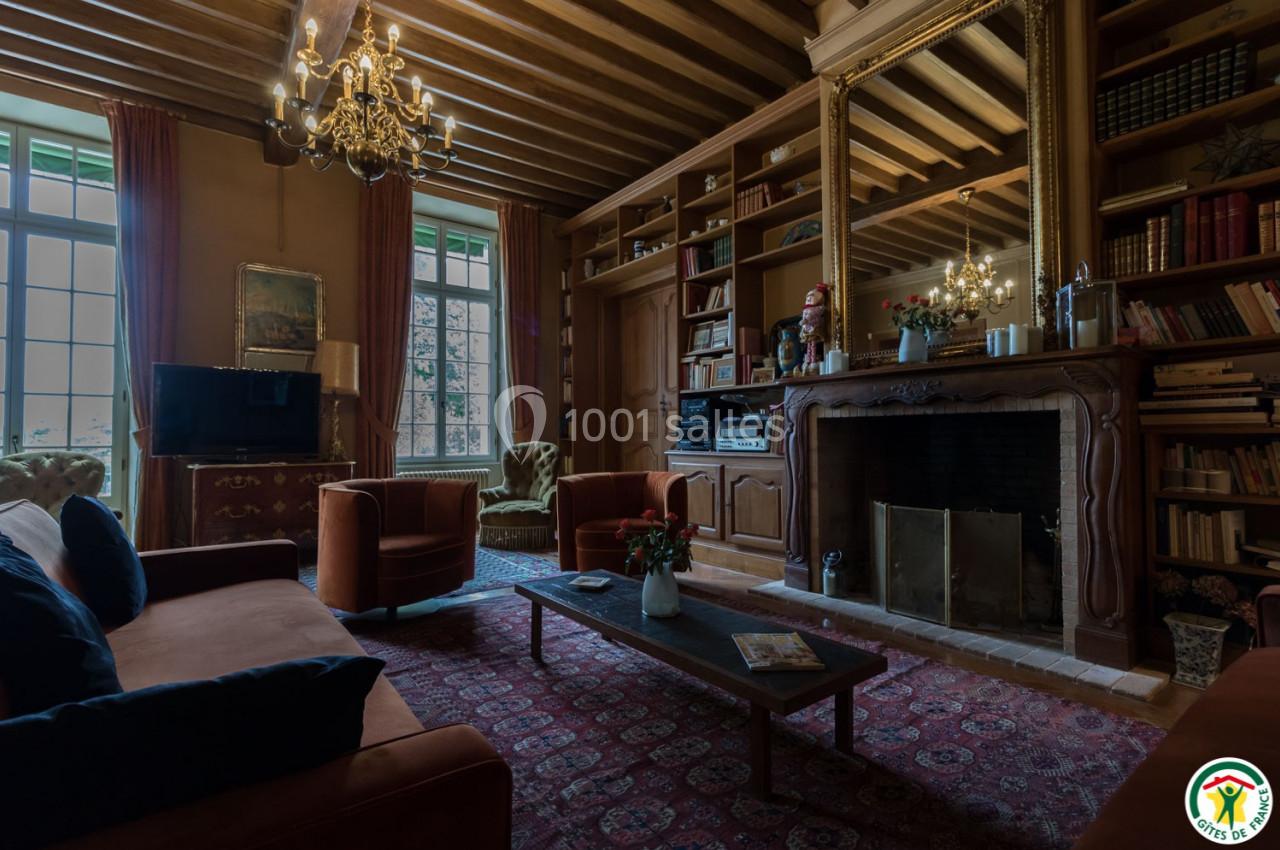 Salon chaleureux avec cheminée, mobilier ancien, bibliothèque en bois, lustres et grandes fenêtres donnant sur l'extérieur.