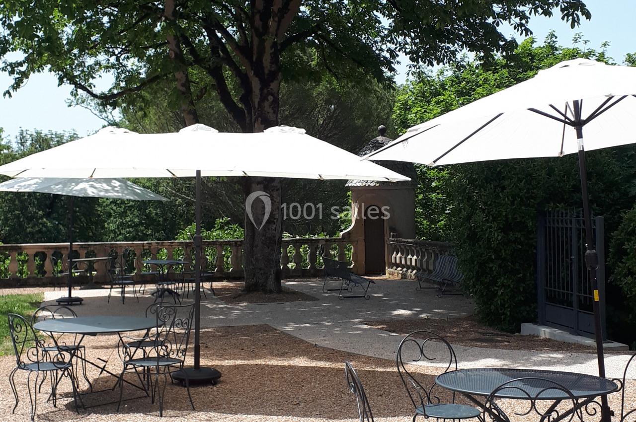 Terrasse en gravier avec tables et chaises en fer forgé, ombragée par des parasols et un grand arbre.