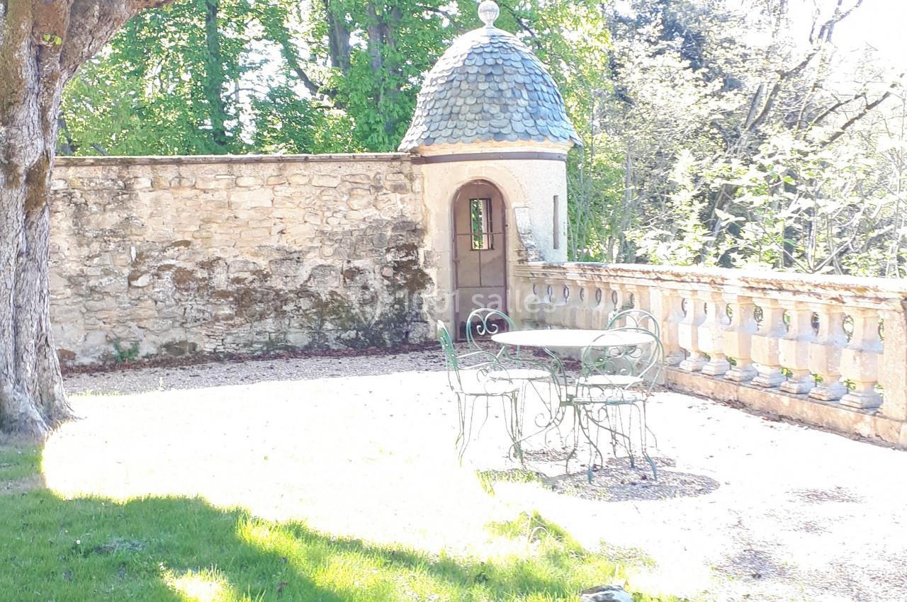 Terrasse ensoleillée avec table et chaises en fer forgé, bordée d'un mur en pierre et d'un petit pavillon.