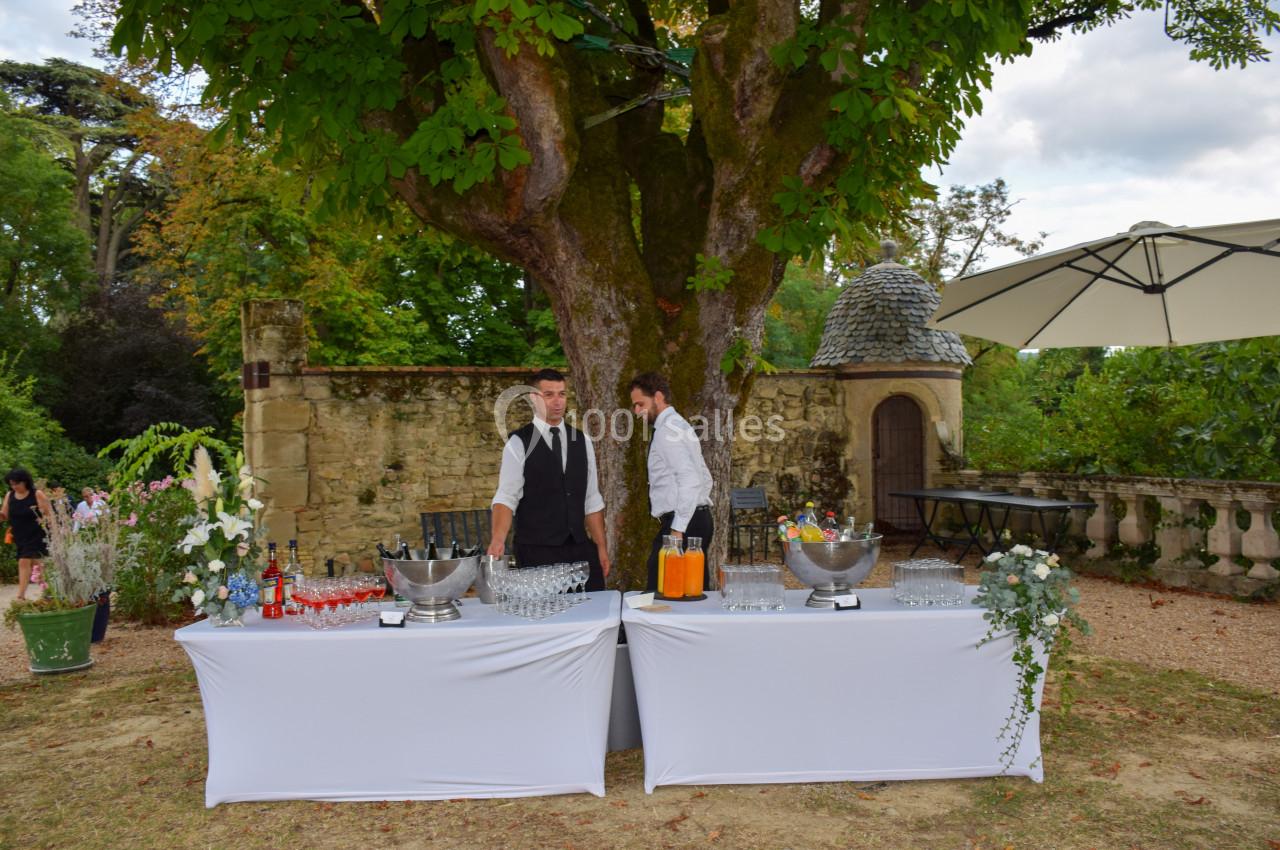 Deux serveurs derrière une table dressée pour un buffet en extérieur, sous un grand arbre dans un cadre champêtre.