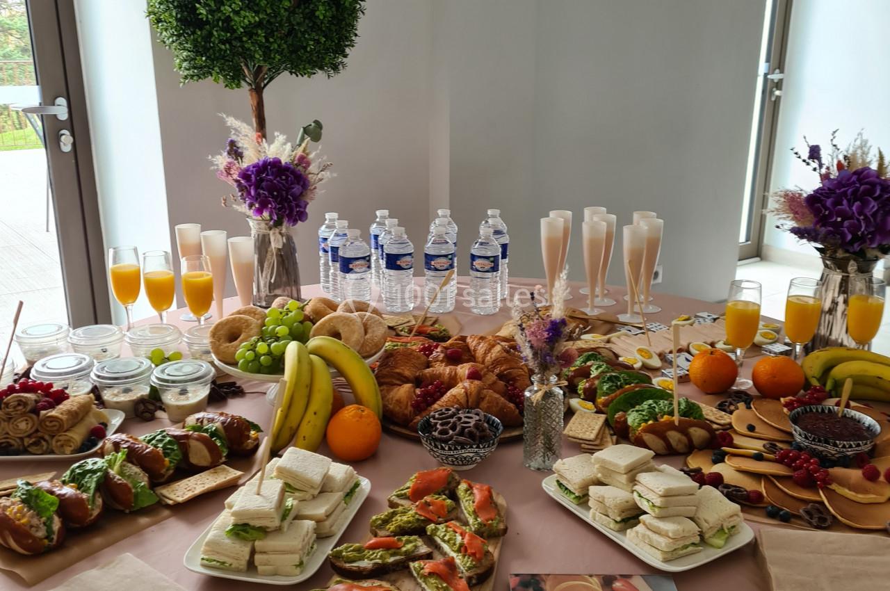 Table garnie de viennoiseries, fruits, sandwichs, boissons et décorations florales dans un cadre lumineux.