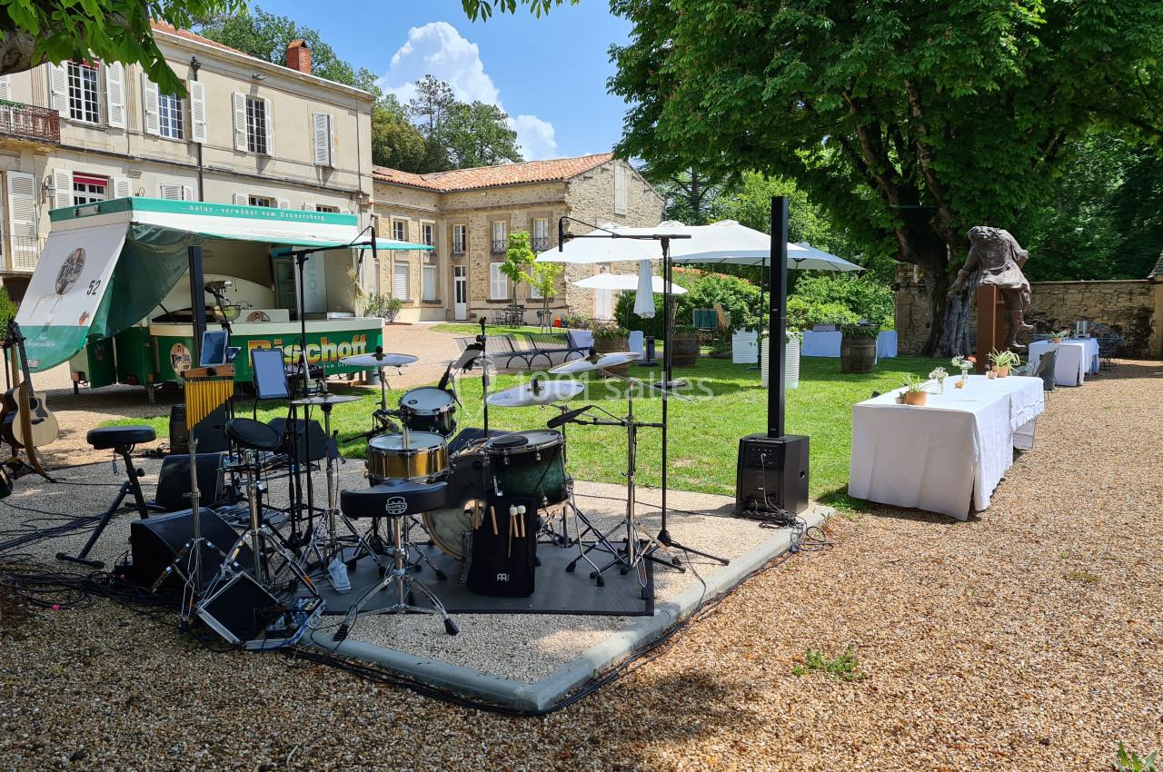 Scène extérieure avec batterie installée, food truck et tables dressées dans un jardin devant un bâtiment ancien.