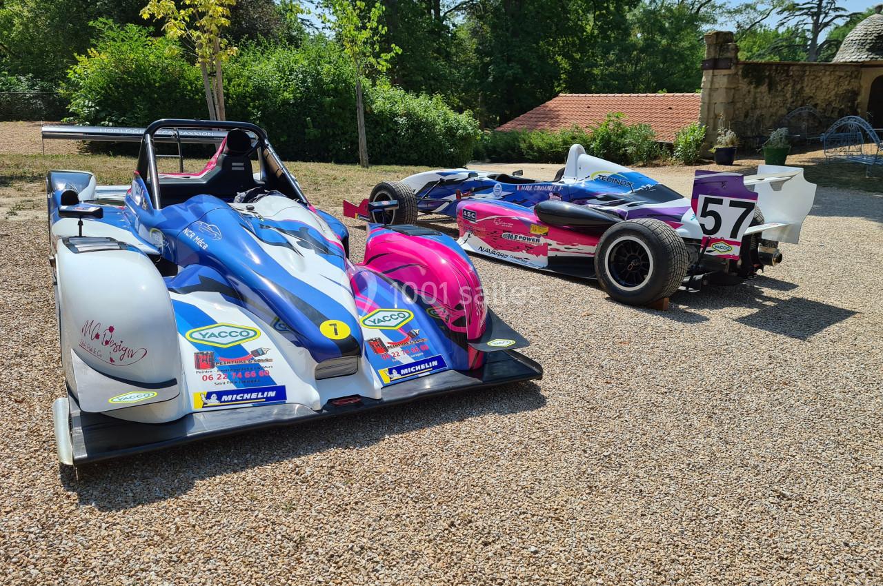 Deux voitures de course colorées stationnées sur un sol gravillonné, entourées de végétation et de bâtiments.