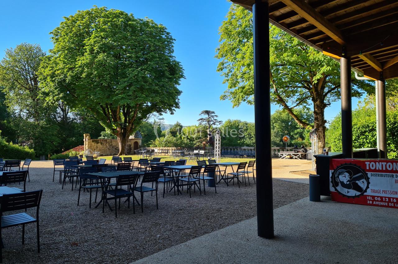Terrasse extérieure avec tables et chaises vides, entourée d'arbres et d'un espace vert sous un ciel dégagé.