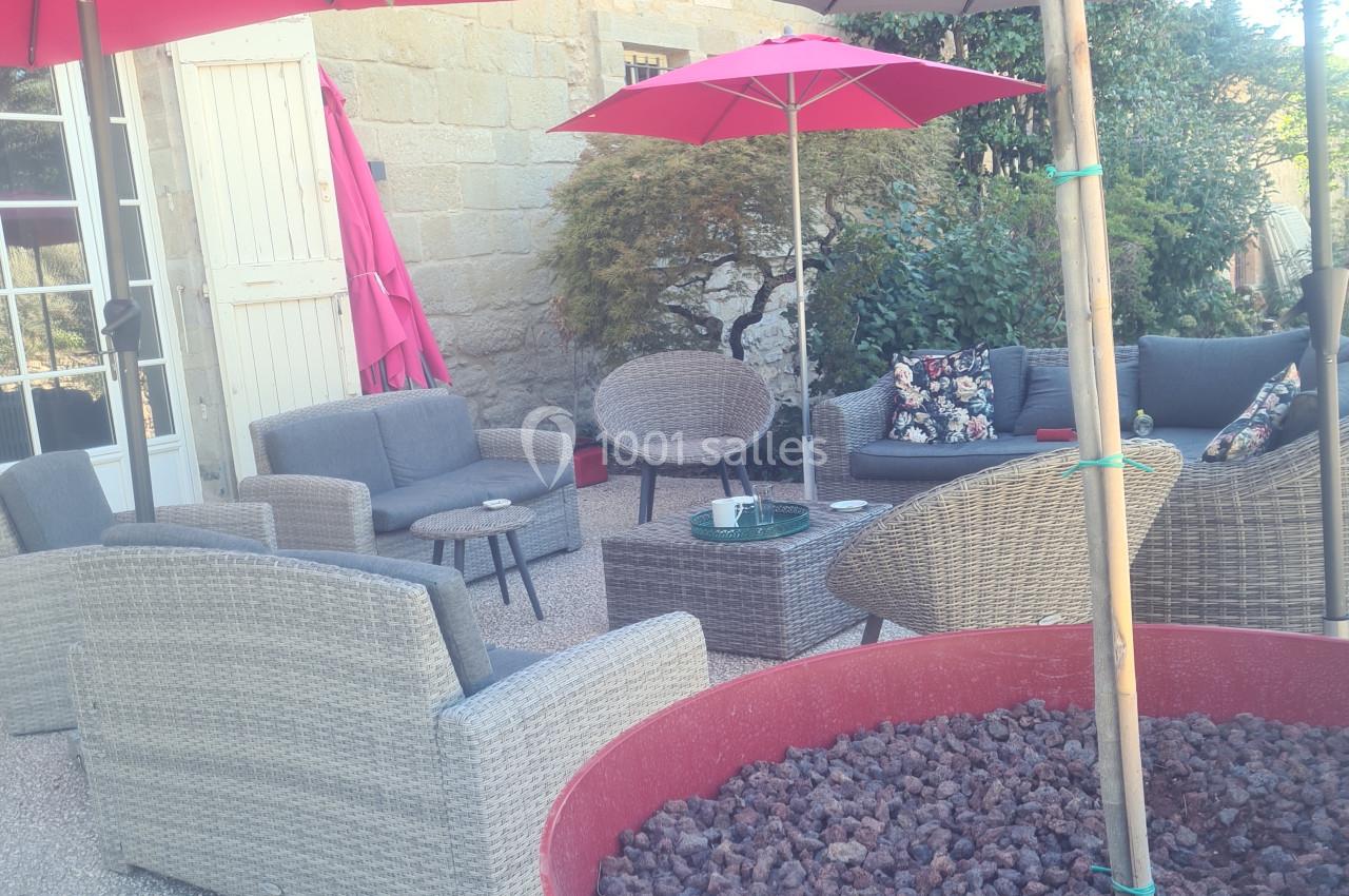 Salon de jardin avec fauteuils en rotin, parasols colorés et grande jardinière rouge sur une terrasse ensoleillée.
