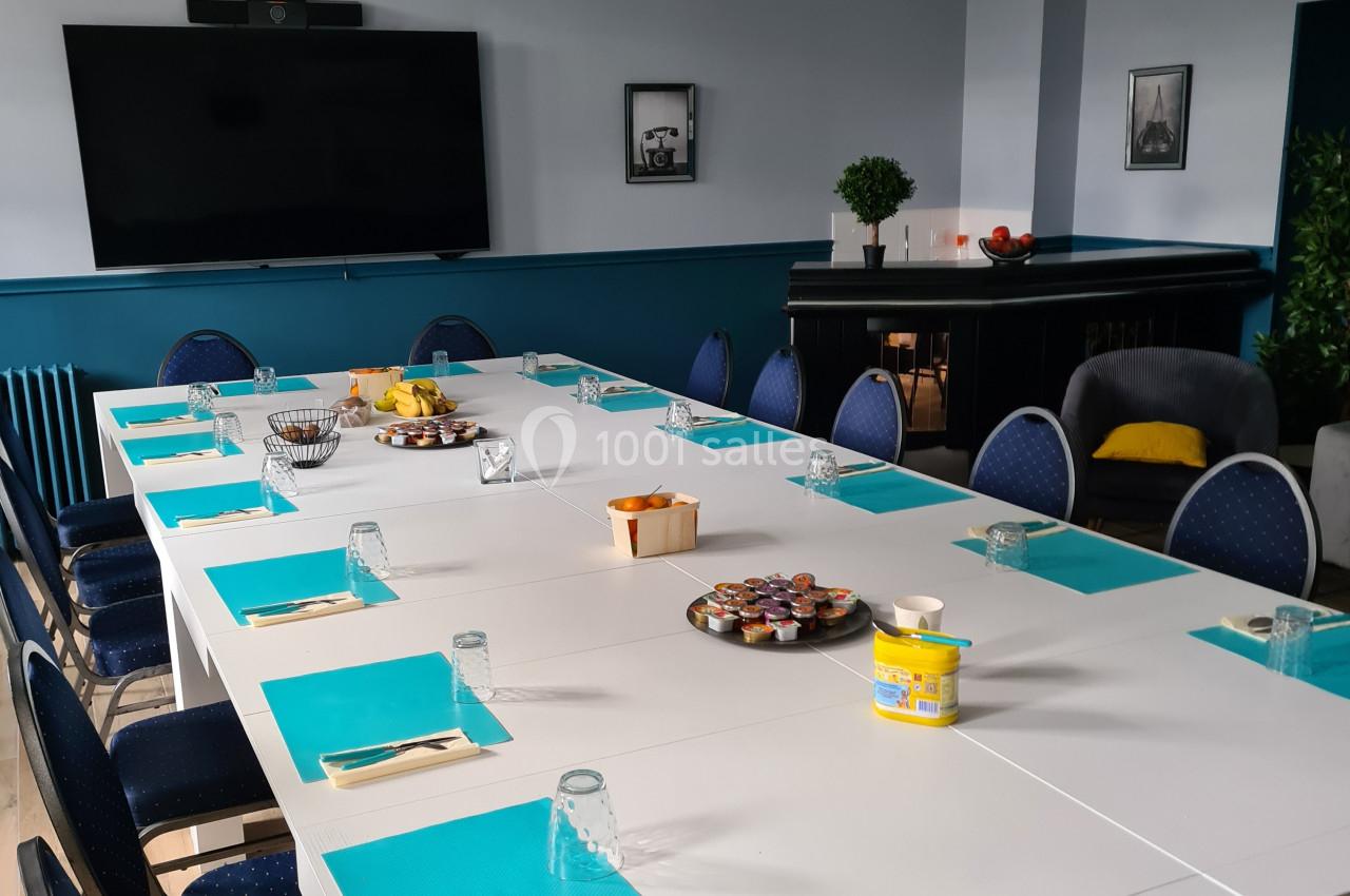 Salle de réunion avec une grande table blanche, sets de table bleus, chaises, écran mural et collations disposées.