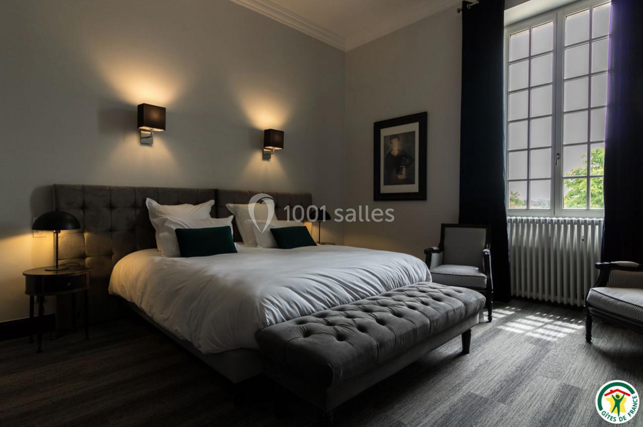 Chambre élégante avec lit double, éclairage tamisé, fauteuils et grande fenêtre laissant entrer la lumière naturelle.