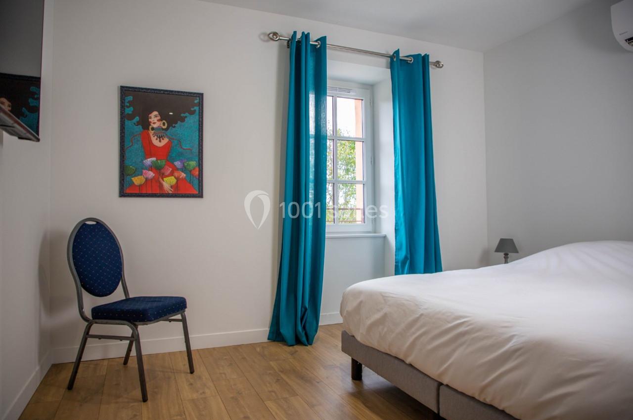 Chambre lumineuse avec lit double, chaise, tableau coloré au mur, rideaux bleus et fenêtre donnant sur un extérieur…