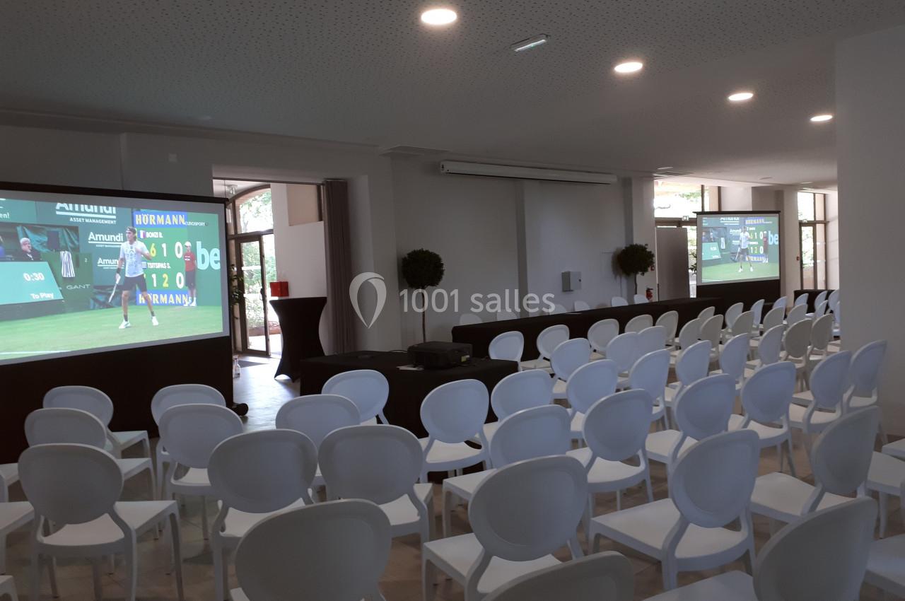 Salle de conférence avec des rangées de chaises blanches et des écrans projetant un match de tennis.