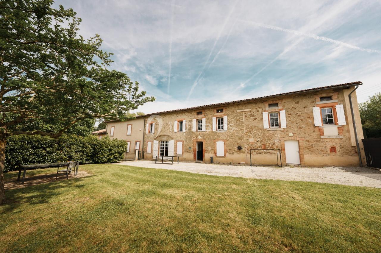 Bâtisse en pierre avec volets blancs, entourée de pelouse et d'arbres, sous un ciel bleu parsemé de traînées d'avion.