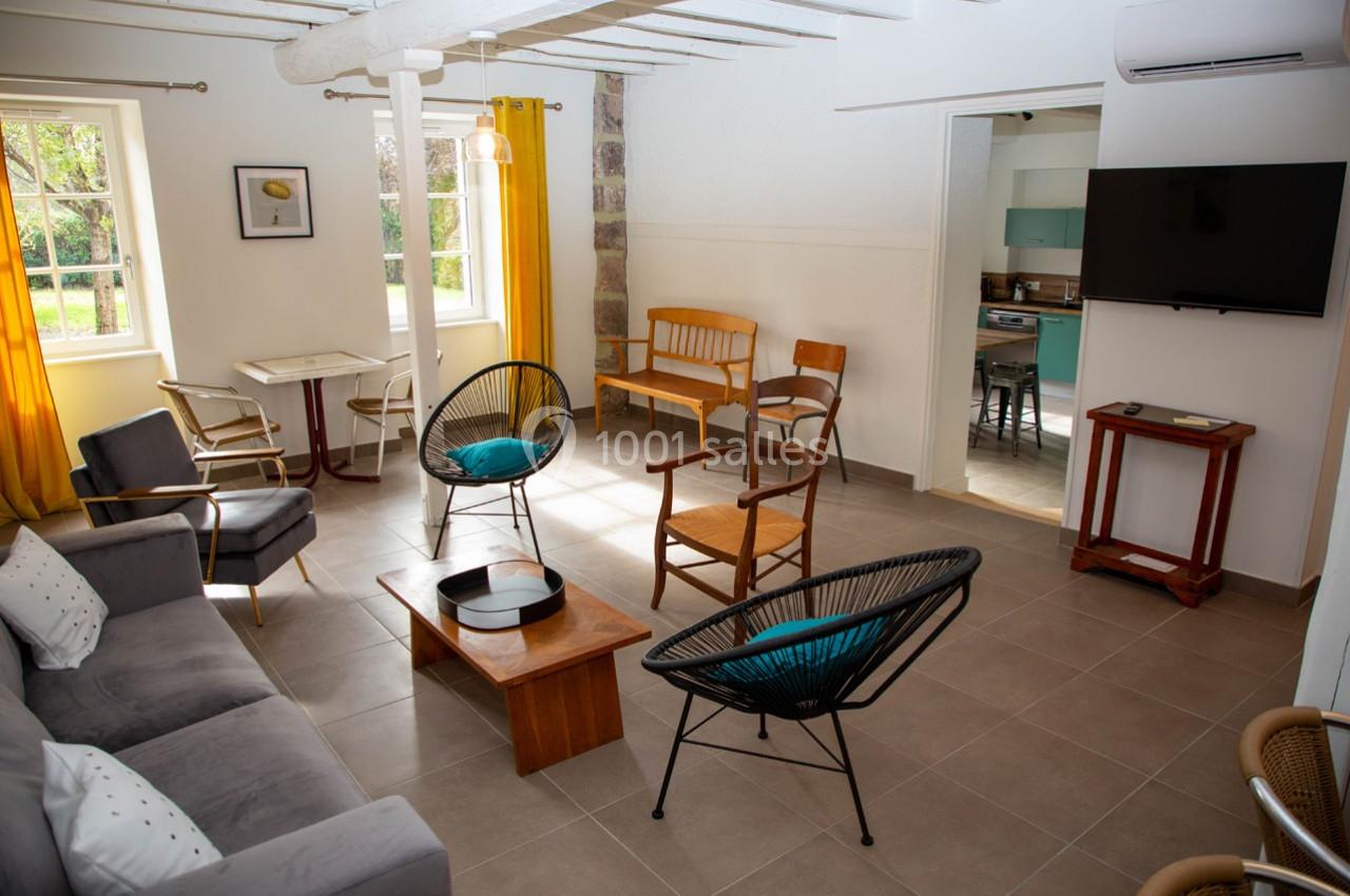 Salon lumineux avec canapé, fauteuils, table basse, télé murale, grandes fenêtres et vue sur une cuisine ouverte.