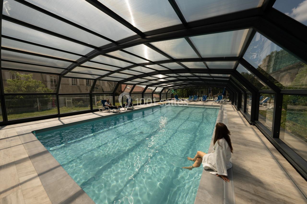 Piscine couverte avec structure vitrée, une femme assise au bord enveloppée dans une serviette.
