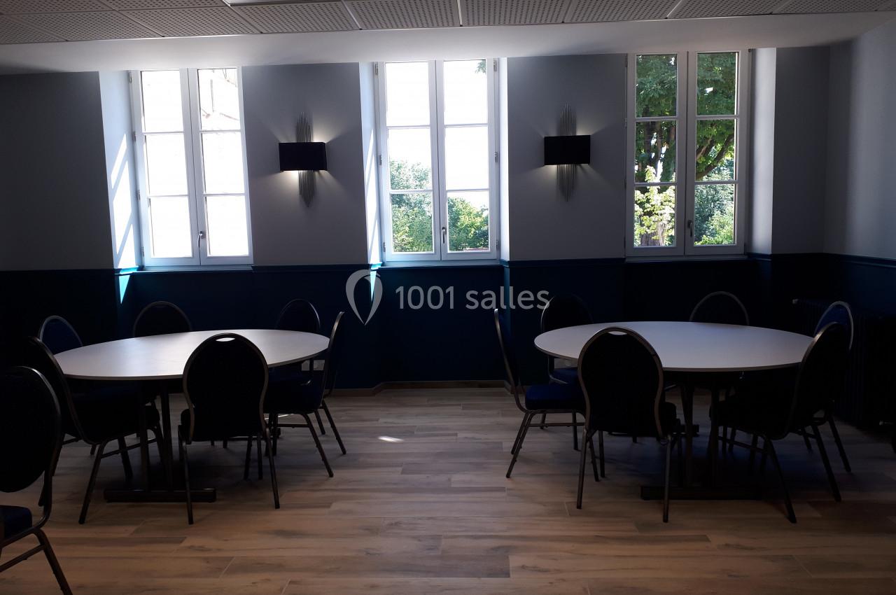 Salle lumineuse avec parquet, tables rondes et chaises noires, éclairée par des fenêtres donnant sur des arbres.