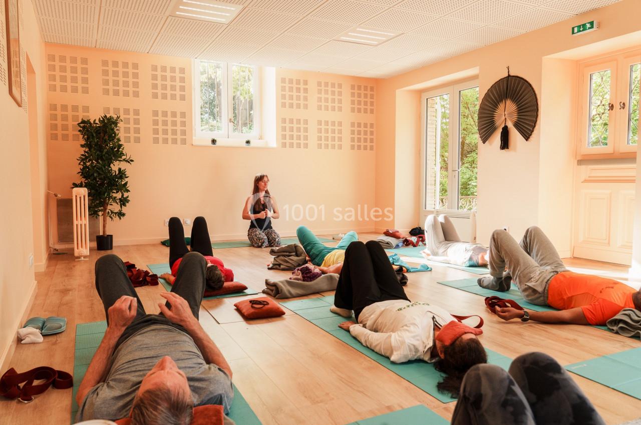 Personnes allongées sur des tapis de yoga dans une salle lumineuse, guidées par une instructrice assise au fond.