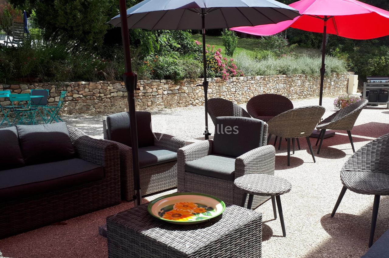 Salon de jardin en rotin avec tables et parasols colorés sur une terrasse ensoleillée entourée de verdure.