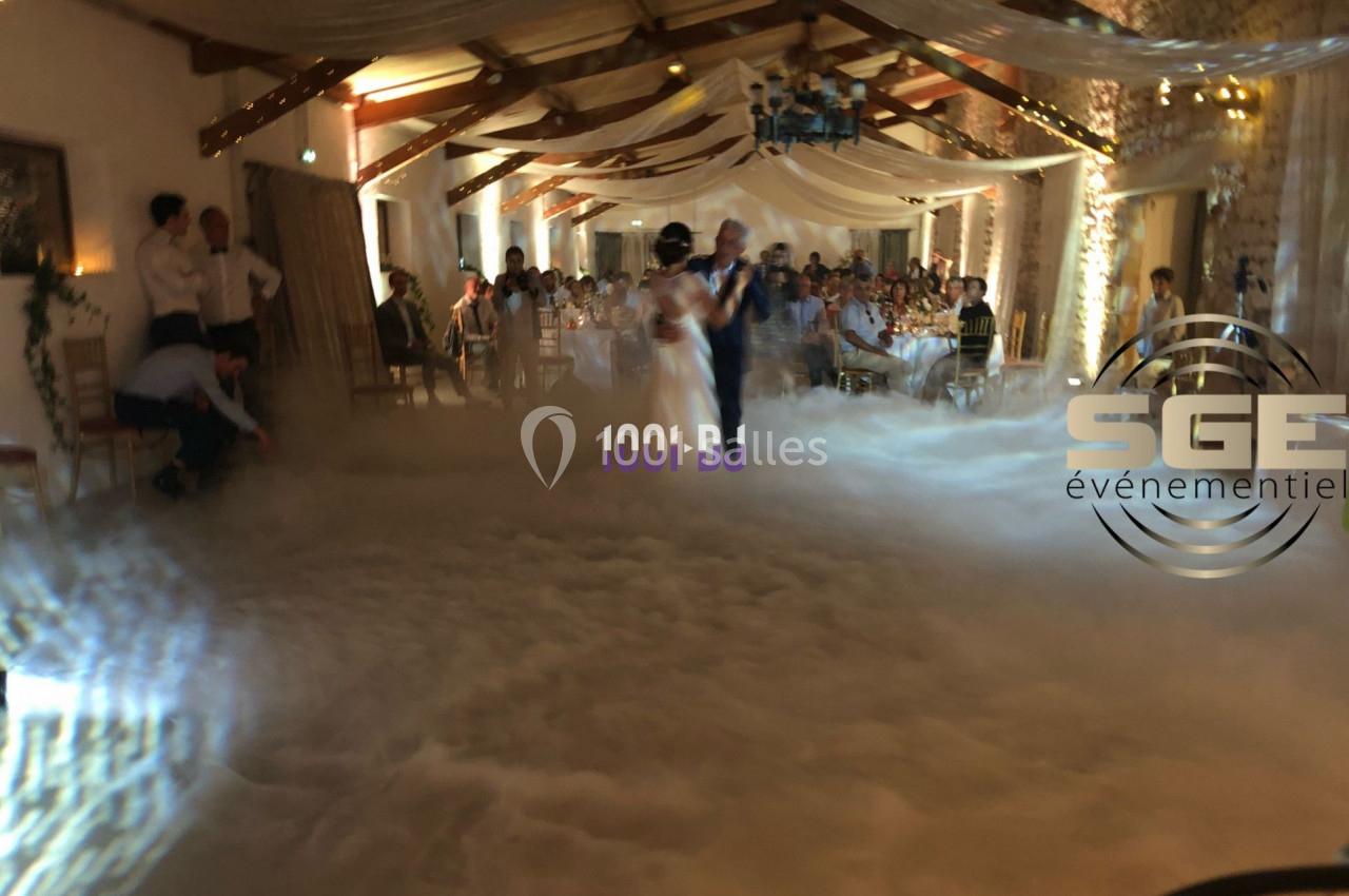 Un couple danse dans une salle de réception décorée, entourée de fumée au sol et d'invités assis.