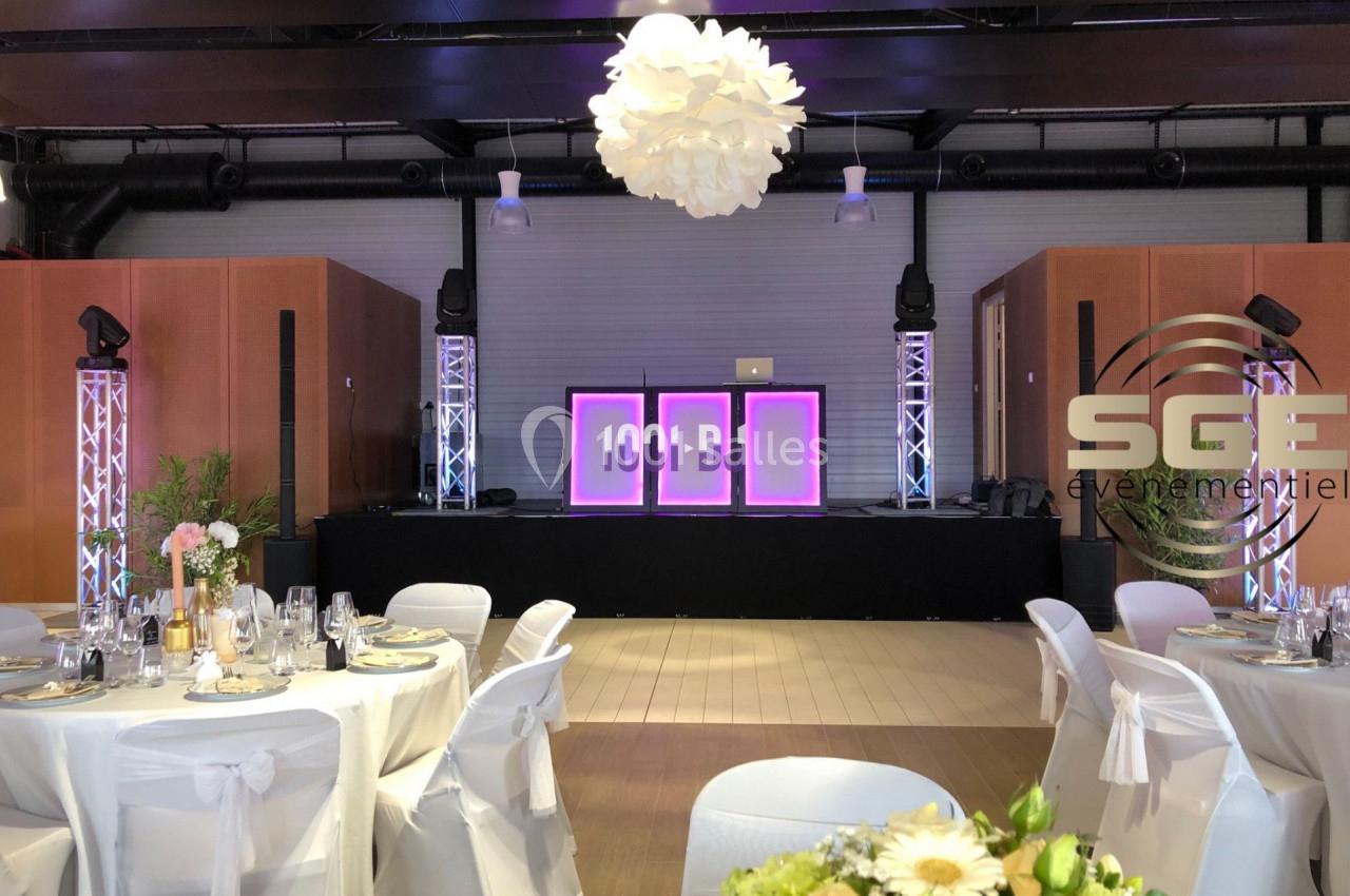 Salle décorée pour un événement avec tables dressées, chaises blanches et scène équipée de lumières et écrans lumineux.