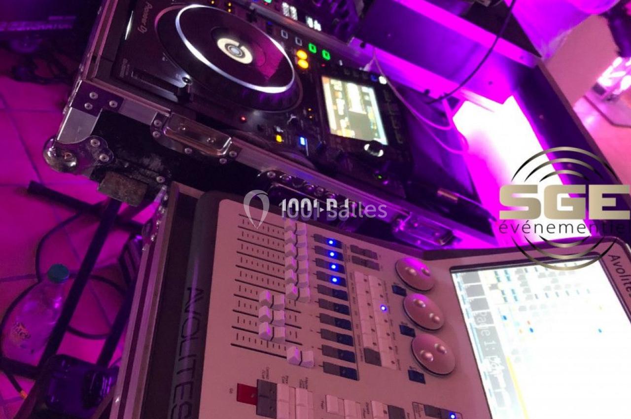 Matériel de DJ et console d'éclairage sur une table, entourés d'équipements techniques dans une ambiance lumineuse violette.