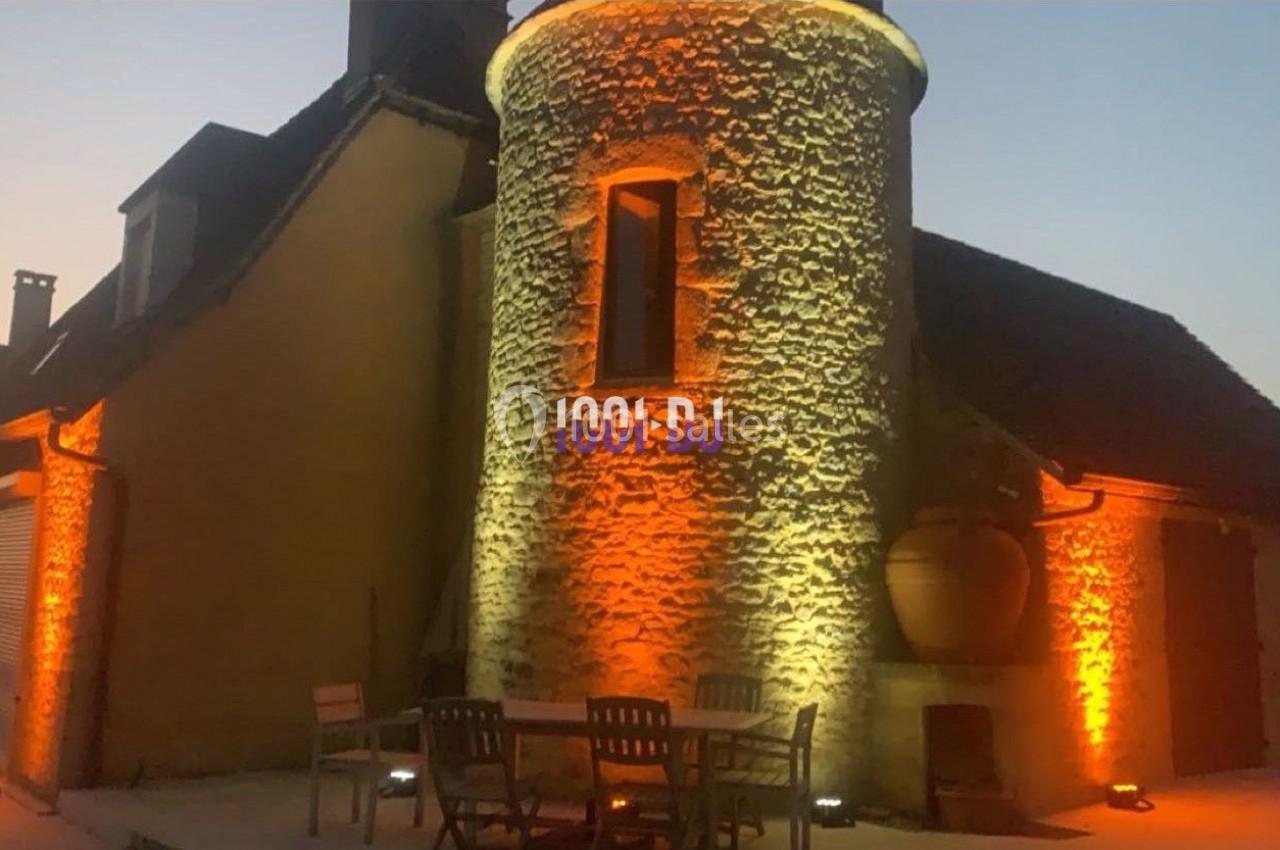 Maison en pierre avec une tour éclairée par des lumières orange au crépuscule, table et chaises au premier plan.