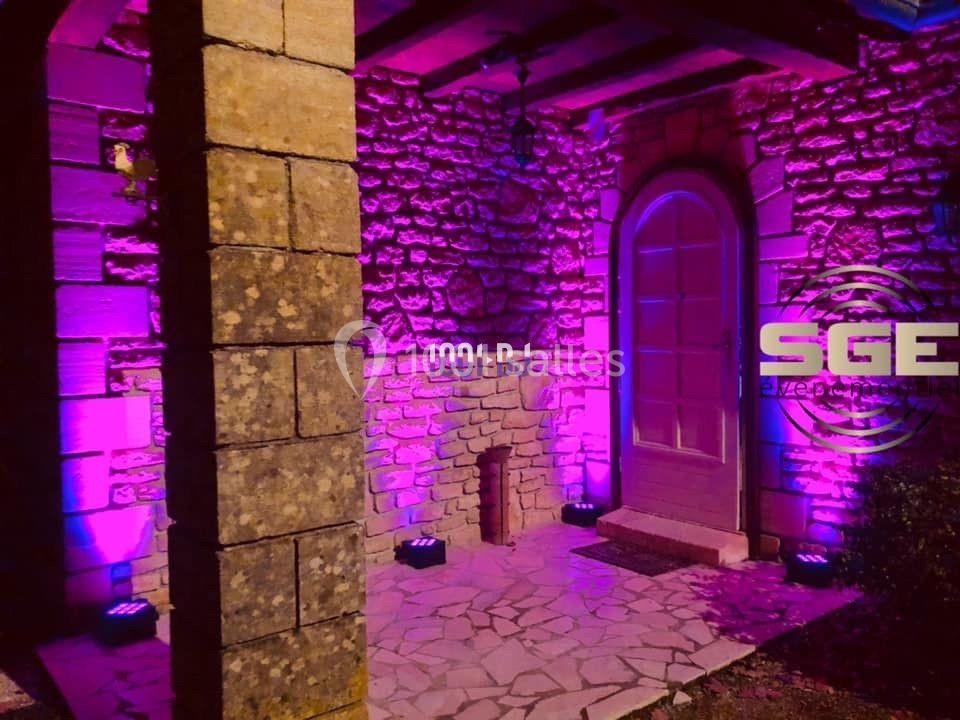 Façade en pierre éclairée par des lumières violettes, avec une porte voûtée et un sol en dalles.