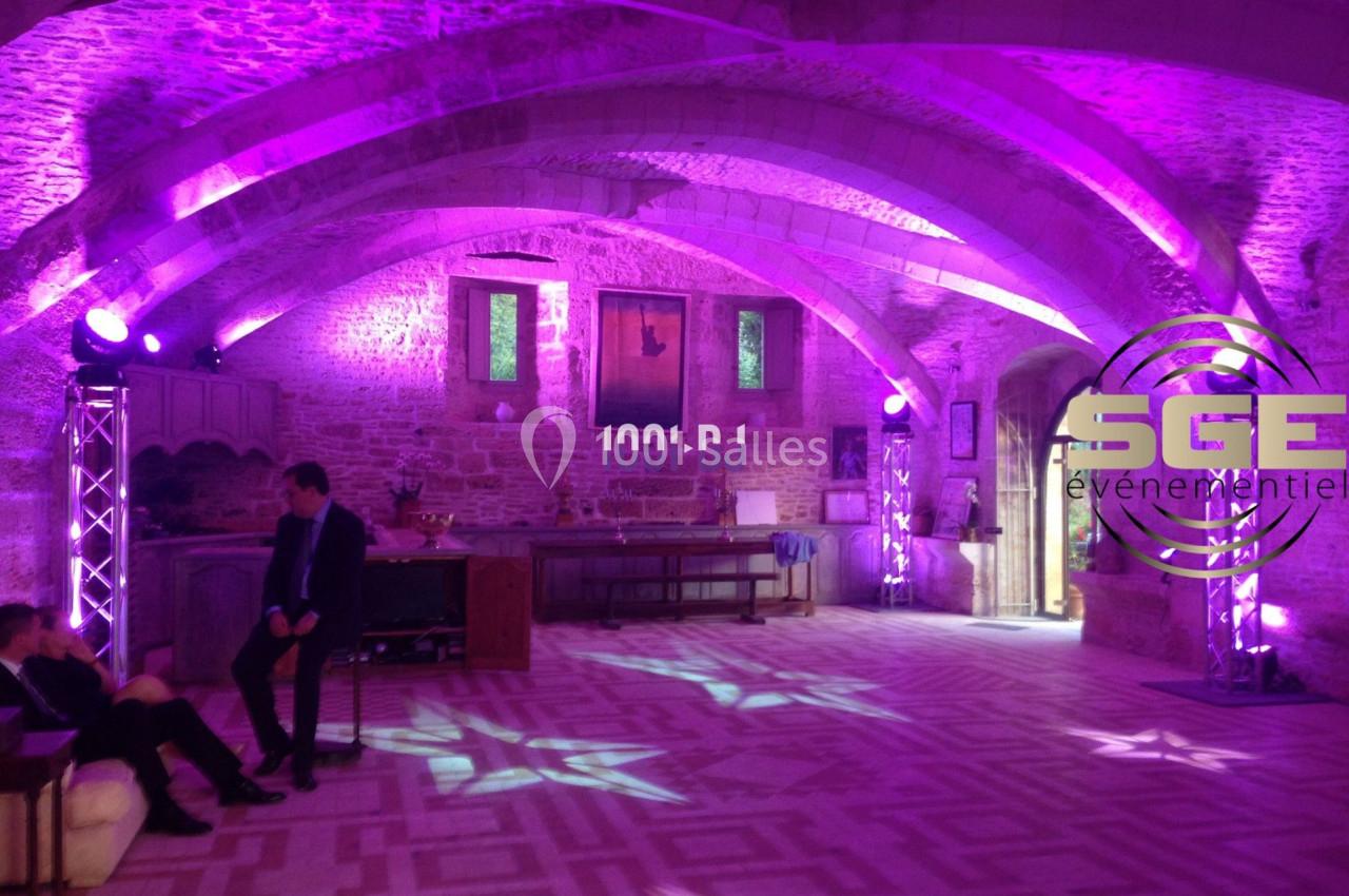 Salle voûtée en pierre éclairée par des lumières violettes, avec mobilier ancien et décorations murales.