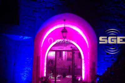 Façade en pierre éclairée par des lumières violettes, avec une porte voûtée et un sol en dalles.