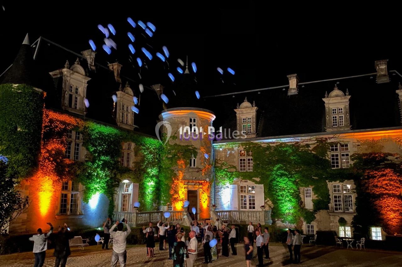 Des personnes libèrent des ballons lumineux devant un château éclairé de nuit par des projecteurs colorés.
