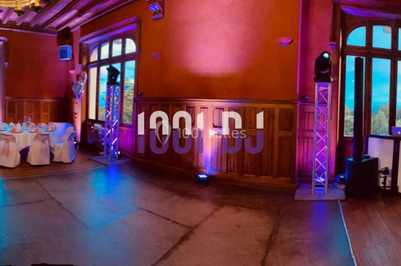 Salle de réception décorée avec des tables rondes, nappes blanches, éclairage coloré et grandes fenêtres.