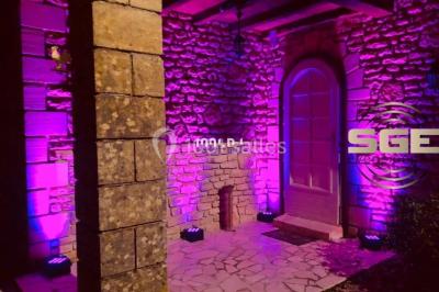 Façade en pierre éclairée par des lumières violettes, avec une porte voûtée et un sol en dalles.