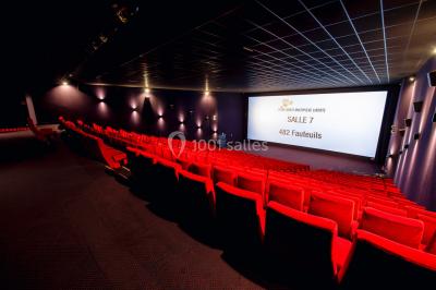 Façade vitrée d'un cinéma moderne sous un ciel bleu, avec trottoir et bâtiments en arrière-plan.