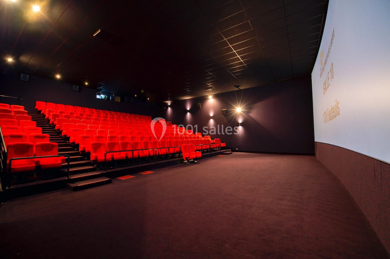Salle de cinéma vide avec des rangées de sièges rouges et un écran blanc illuminé sur la droite.