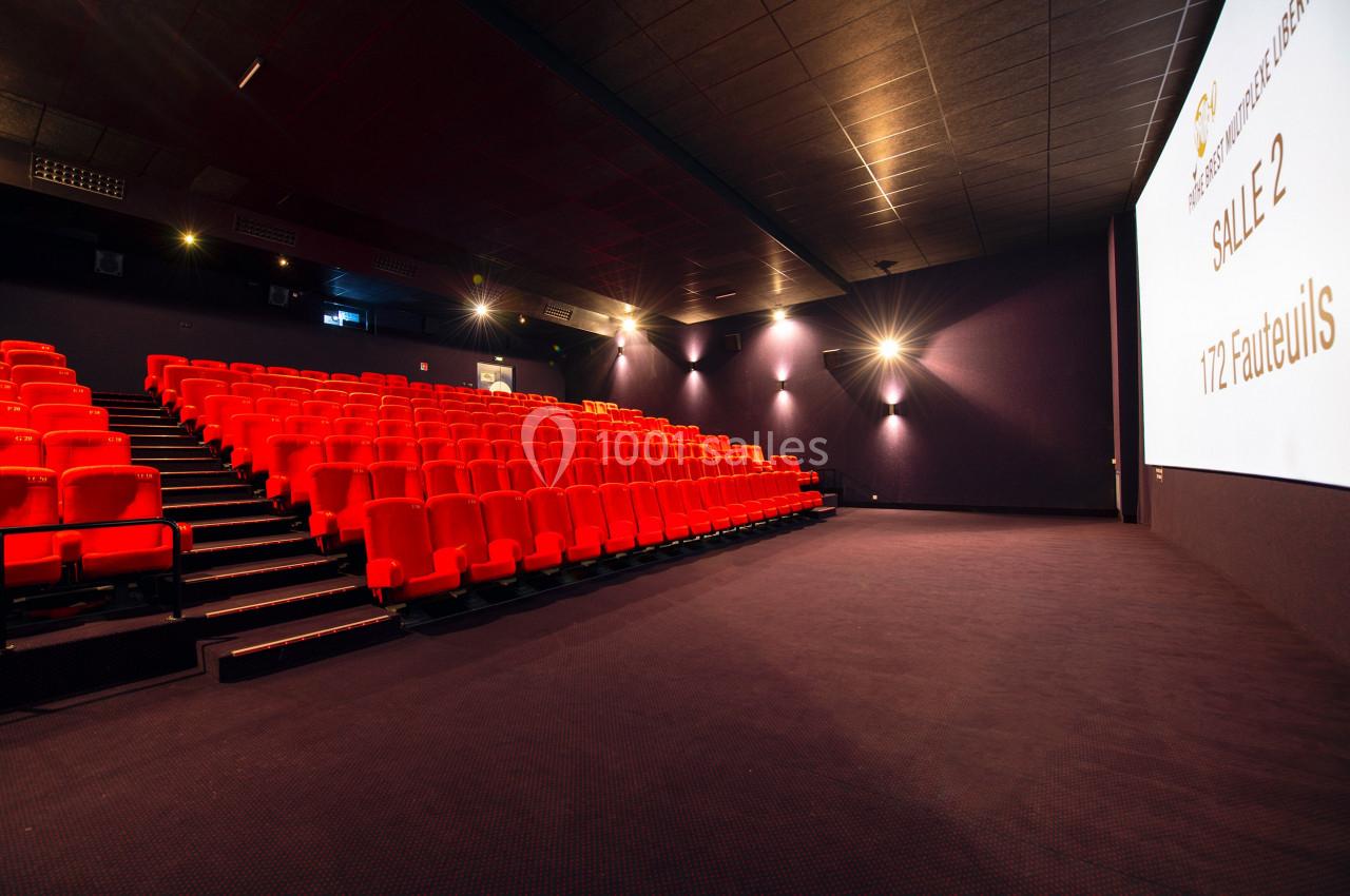 Salle de cinéma vide avec rangées de fauteuils rouges, écran blanc et éclairage tamisé.