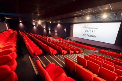 Façade vitrée d'un cinéma moderne sous un ciel bleu, avec trottoir et bâtiments en arrière-plan.