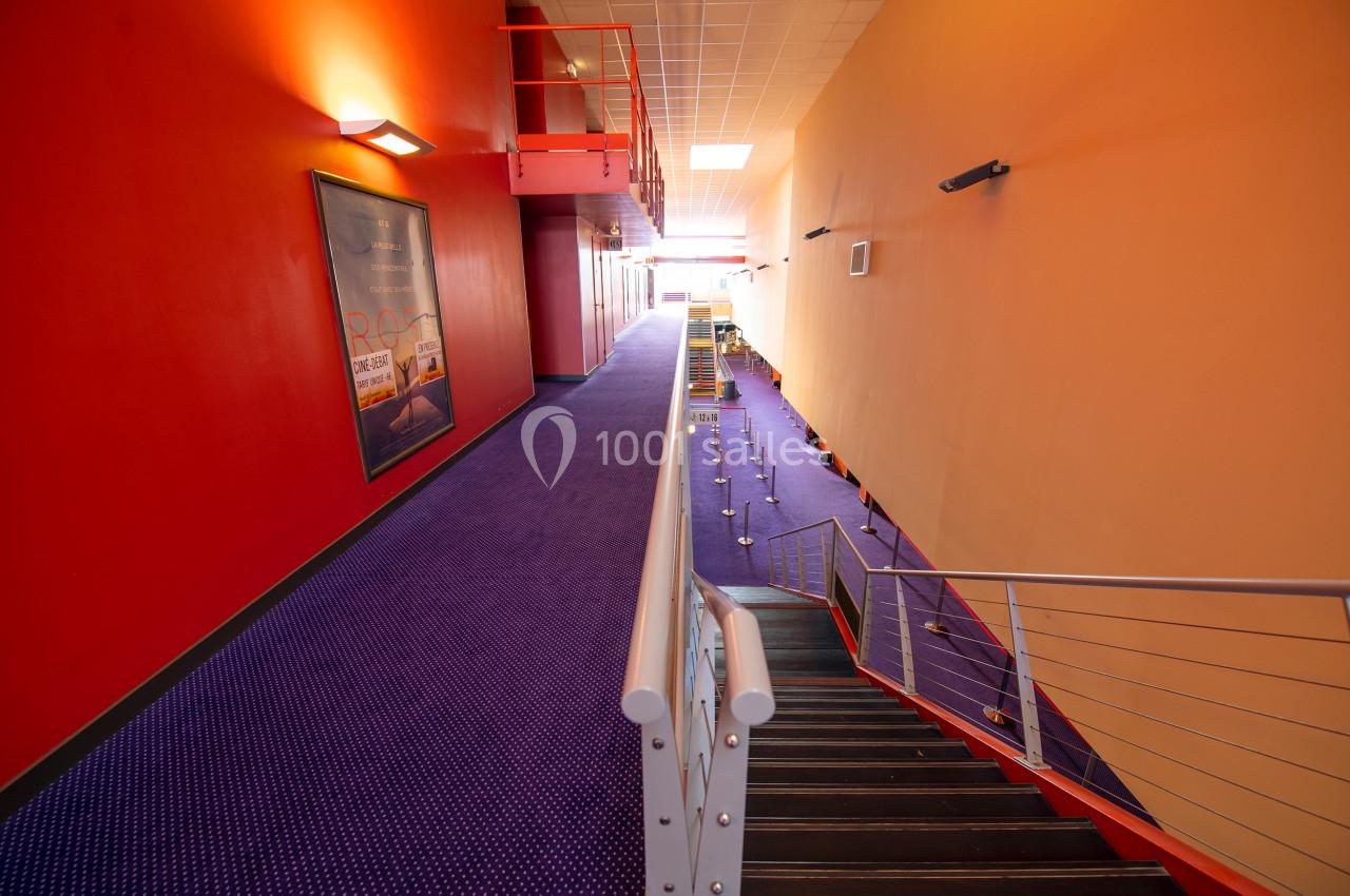 Couloir d'un cinéma avec moquette violette, murs rouges et jaunes, escalier en métal et affiches murales.