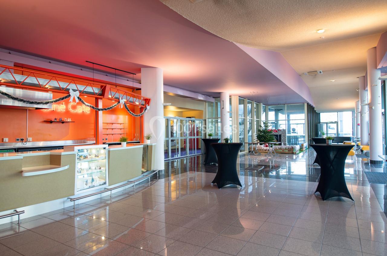 Hall spacieux avec comptoir de café orange, tables hautes noires et grandes baies vitrées laissant entrer la lumière.