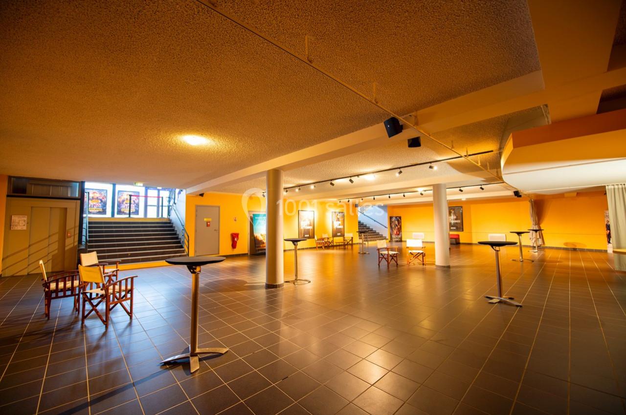 Hall spacieux avec sol carrelé, murs jaunes, tables hautes et chaises, éclairé par des spots au plafond.