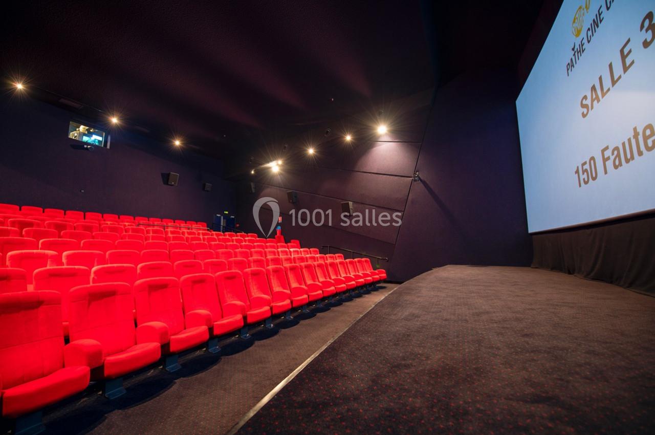 Salle de cinéma vide avec rangées de fauteuils rouges, écran blanc à droite et éclairage tamisé.