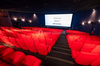 Couloir d'un cinéma avec moquette beige, murs orange, portes numérotées et éclairage chaleureux.