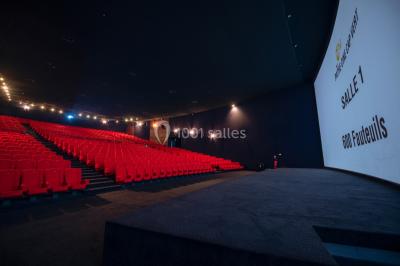 Couloir d'un cinéma avec moquette beige, murs orange, portes numérotées et éclairage chaleureux.