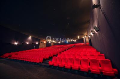 Couloir d'un cinéma avec moquette beige, murs orange, portes numérotées et éclairage chaleureux.