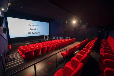 Couloir d'un cinéma avec moquette beige, murs orange, portes numérotées et éclairage chaleureux.
