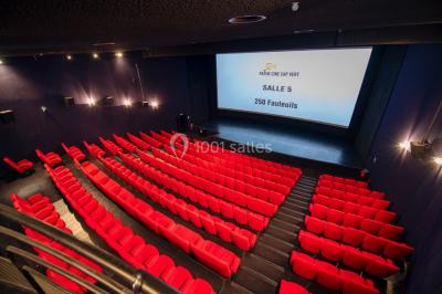 Couloir d'un cinéma avec moquette beige, murs orange, portes numérotées et éclairage chaleureux.