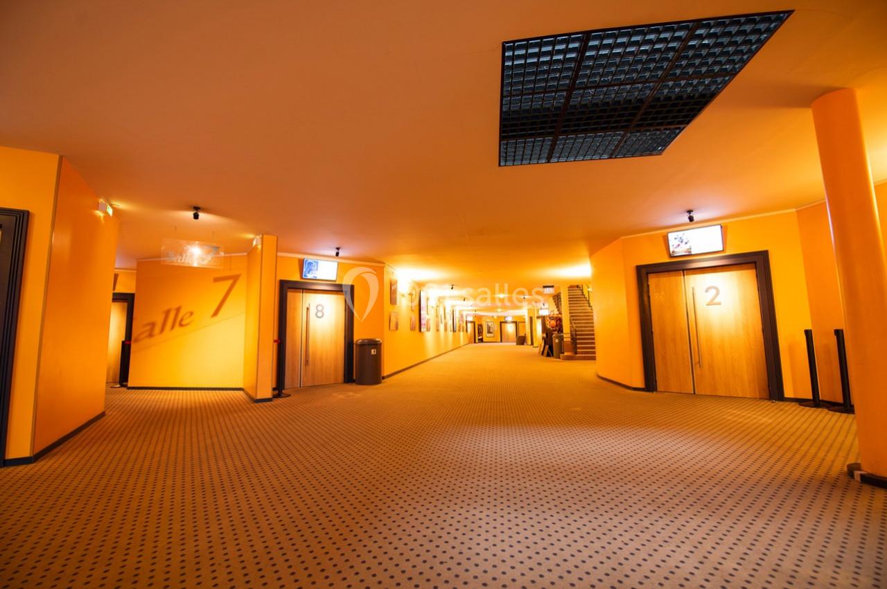 Couloir d'un cinéma avec moquette beige, murs orange, portes numérotées et éclairage chaleureux.