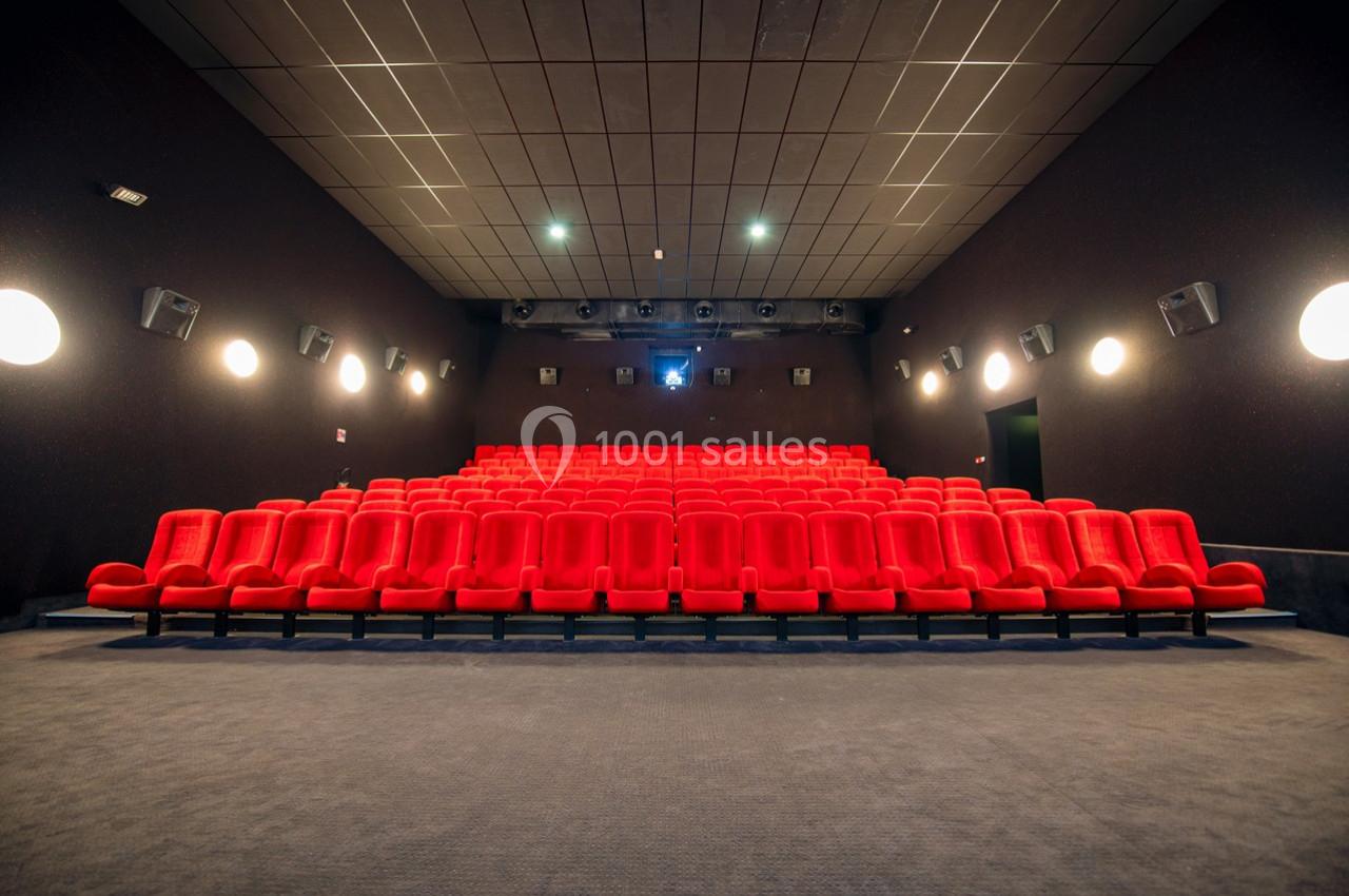 Salle de cinéma vide avec des rangées de fauteuils rouges alignés face à un écran blanc.