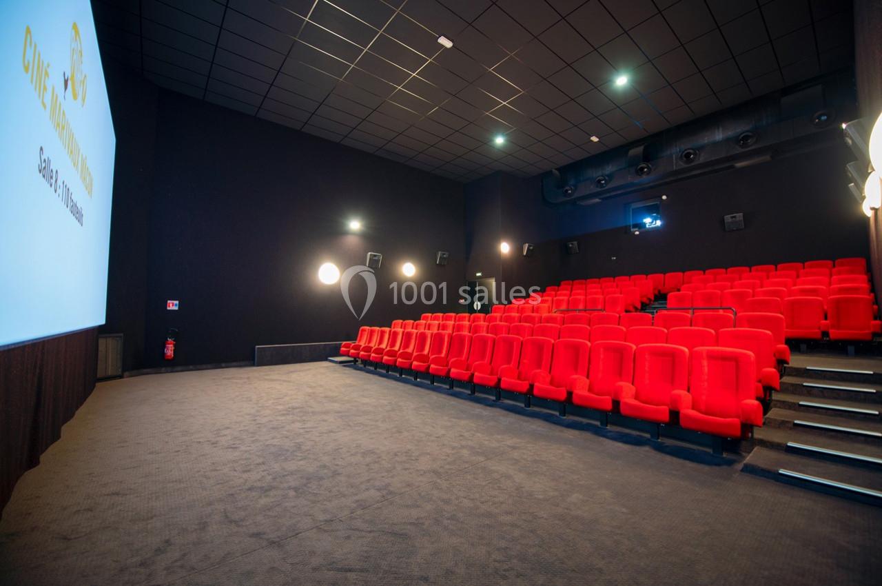 Salle de cinéma vide avec des rangées de sièges rouges, un écran blanc et un éclairage tamisé.