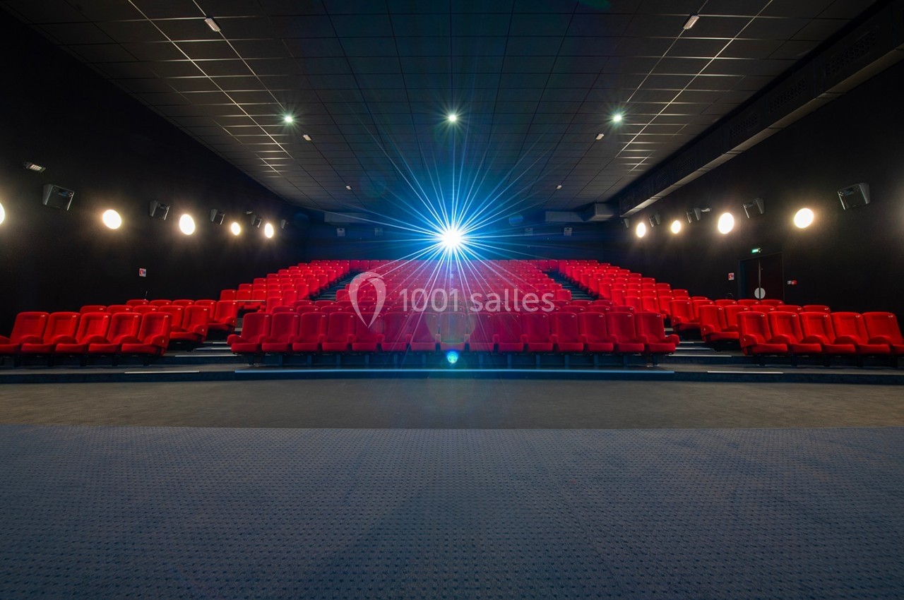 Salle de cinéma vide avec des rangées de sièges rouges et un projecteur allumé au fond.