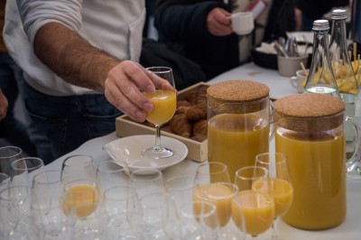 Une personne sert un verre de jus d'orange sur une table garnie de carafes, verres et viennoiseries.