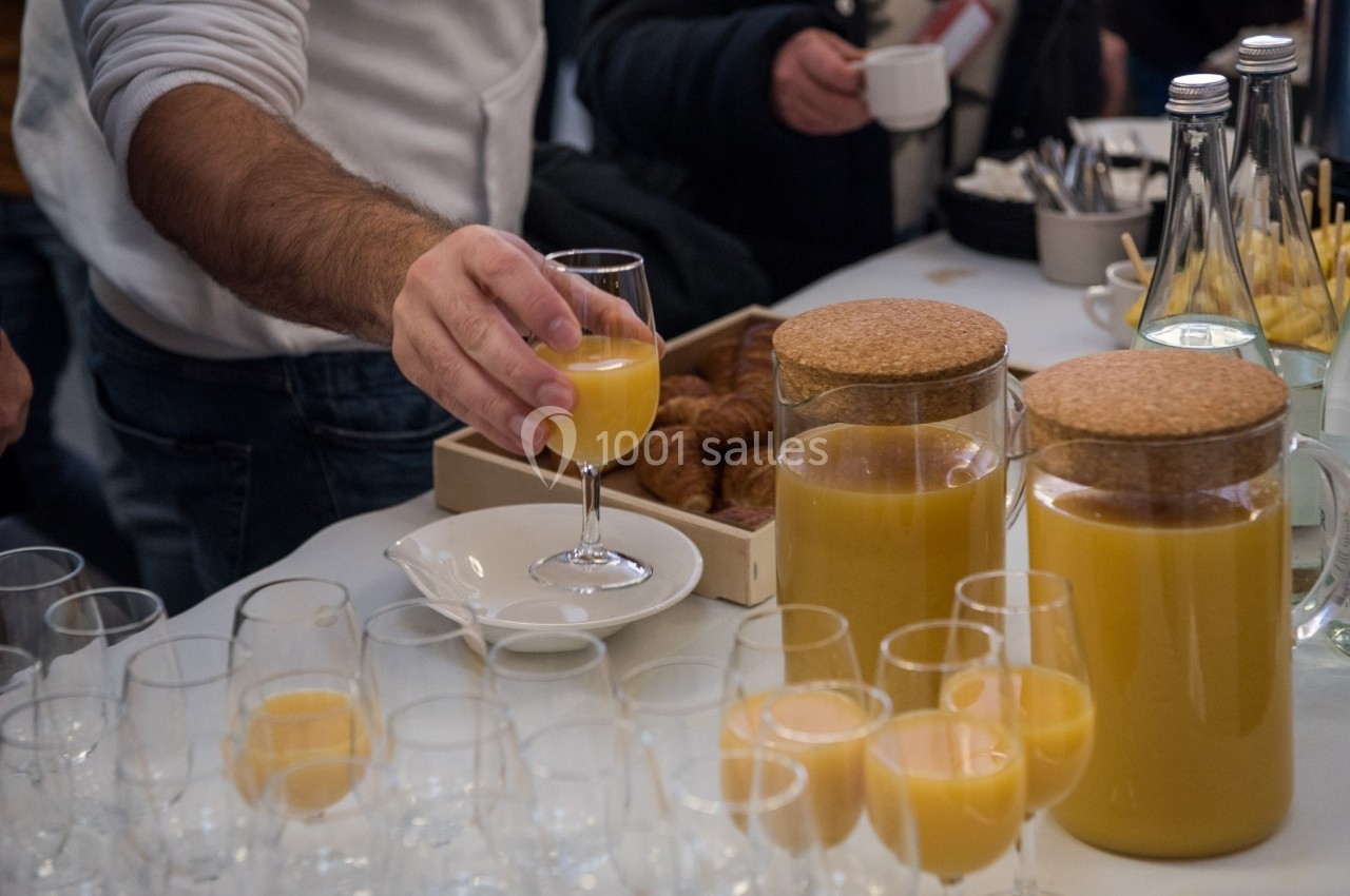 Une personne sert un verre de jus d'orange sur une table garnie de carafes, verres et viennoiseries.