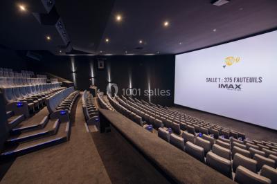 Façade d'un cinéma moderne avec enseigne IMAX, éclairée en soirée, entourée de végétation et d'un parking.
