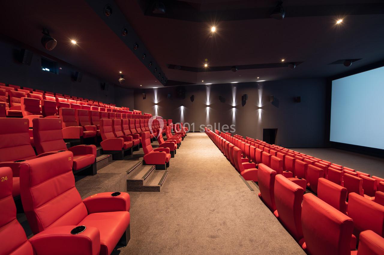 Salle de cinéma vide avec des rangées de fauteuils rouges et un grand écran blanc à droite.